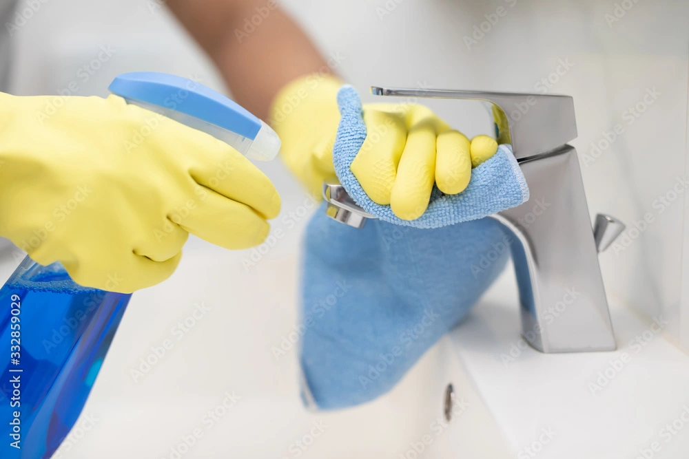Hands wearing gloves cleaning a faucet with a spray bottle and cloth.